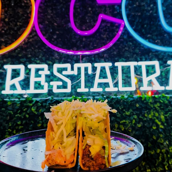 Two hard-shell tacos with ground beef, lettuce, and cheese, in front of a neon Coco Restaurant sign.