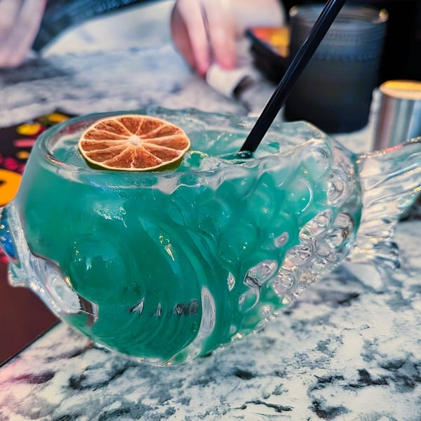 A vibrant blue cocktail in a fish-shaped glass with a dried orange slice and black straw.