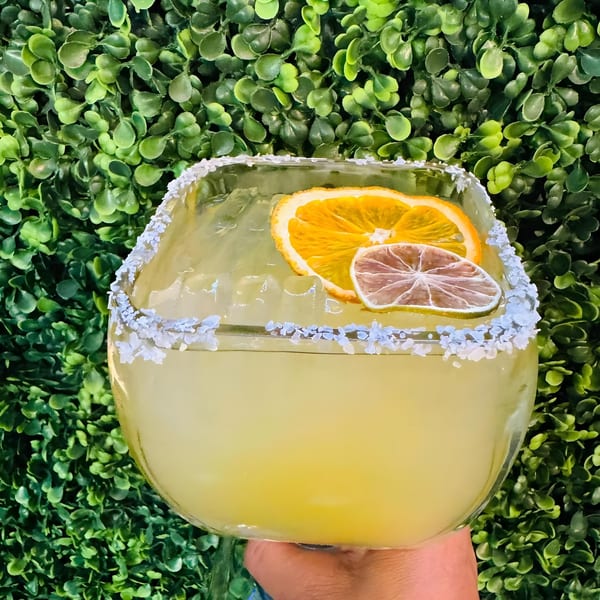 A large Coco Mexican Restaurant margarita with salt rim, orange, and lime slices against a green leafy background.